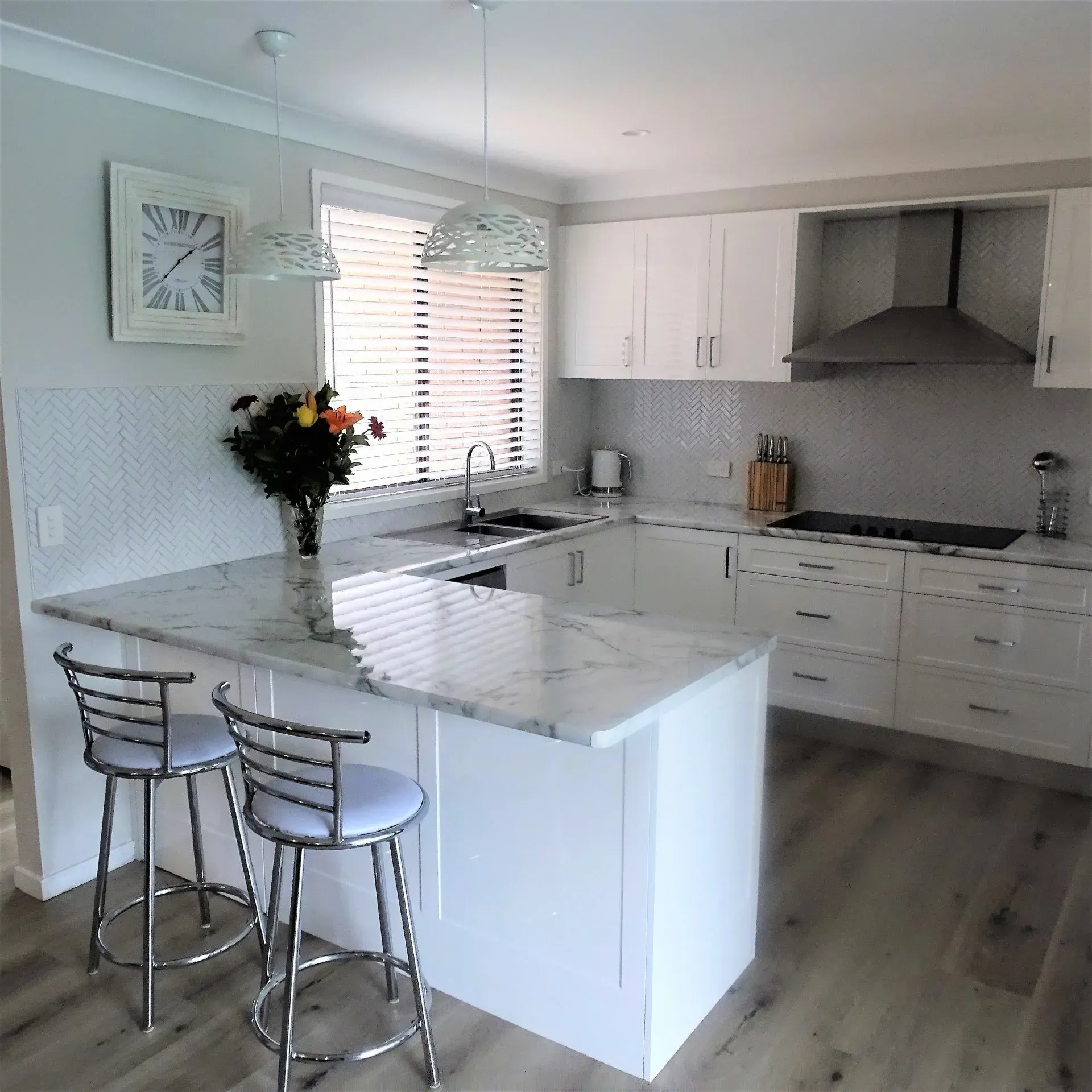 Modern white kitchen with island, marble countertops, stainless steel barstools, and a window.