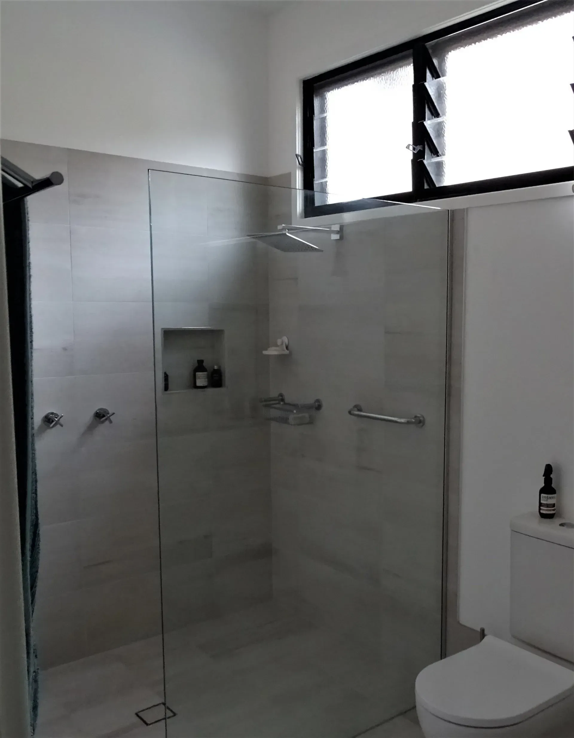 Bathroom with glass shower, grab bar, toilet, and window. White walls, grey tile, and natural light.