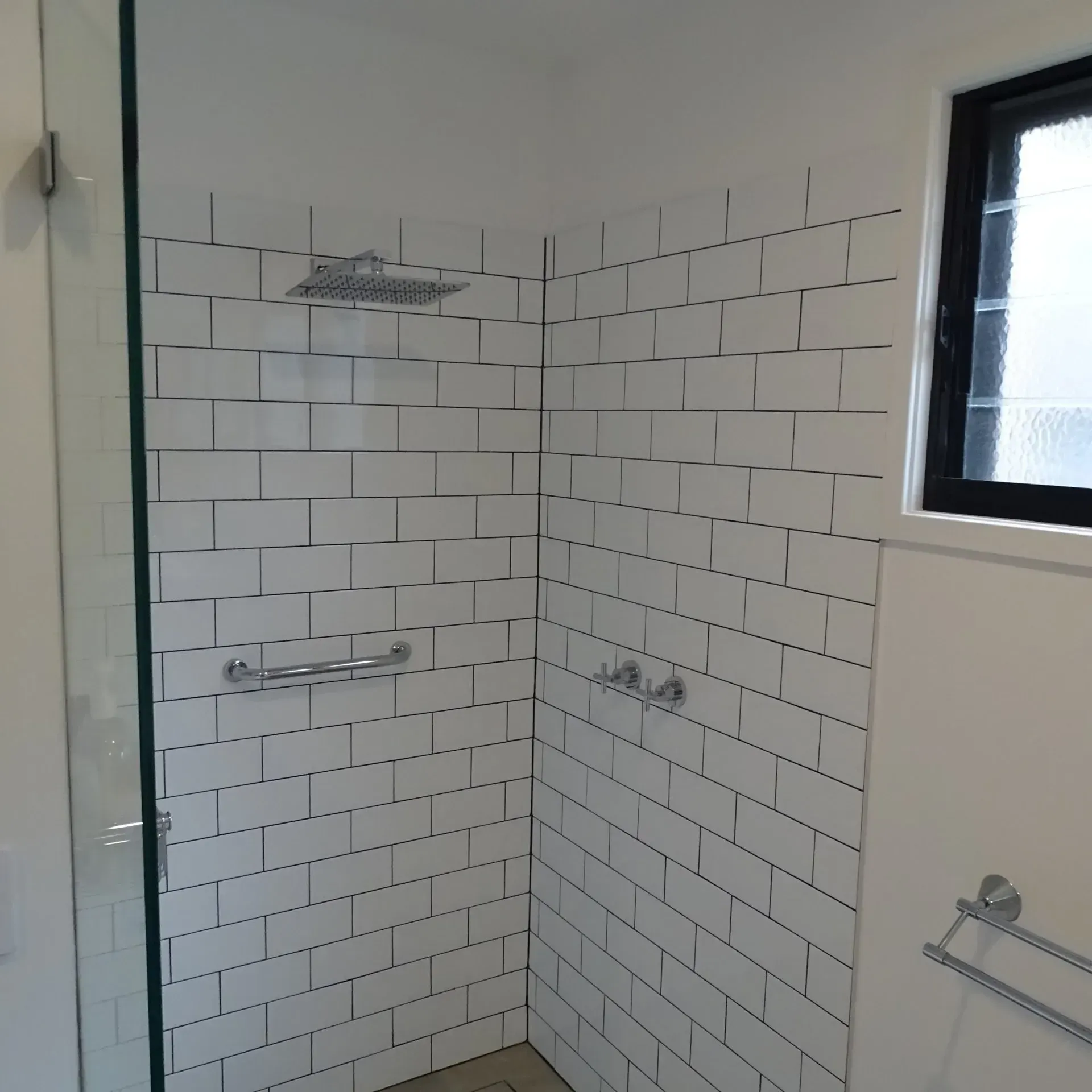 White tiled shower with glass door, rain shower head, and chrome fixtures. Window on the right.