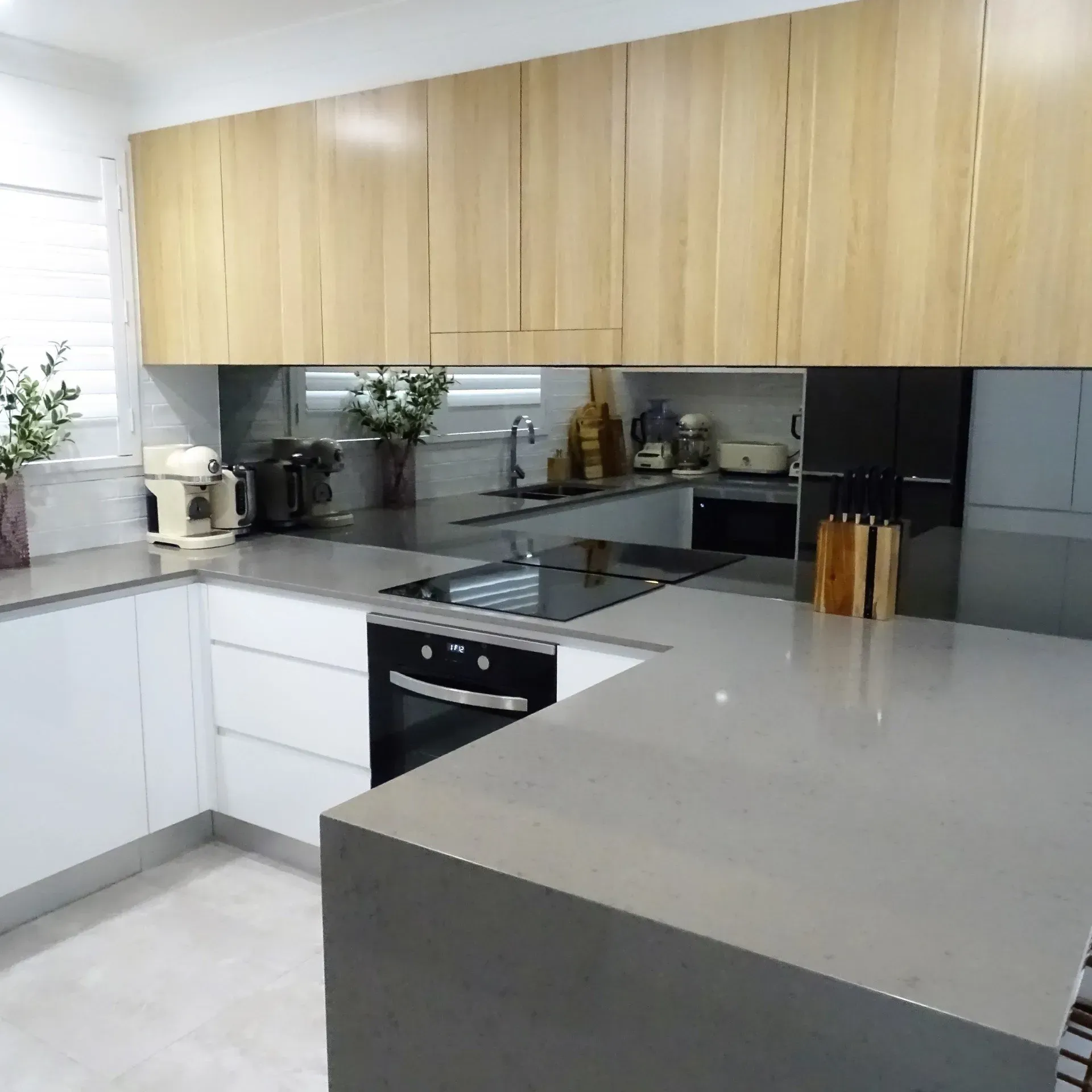 Modern kitchen with light wood upper cabinets, white lower cabinets, gray countertops, and a mirrored backsplash.