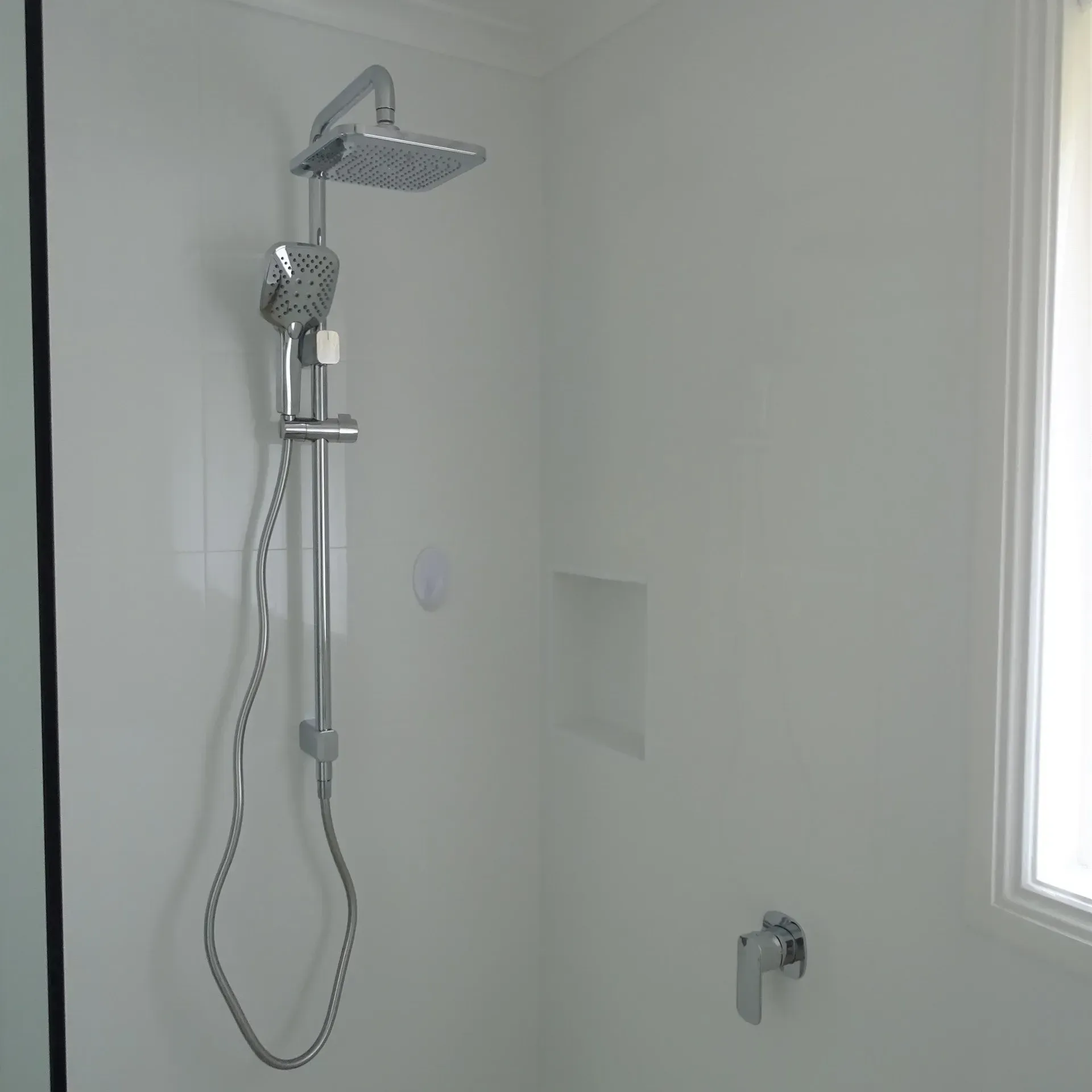 Shower with square showerhead and handheld wand, white walls. Silver faucet and fittings.