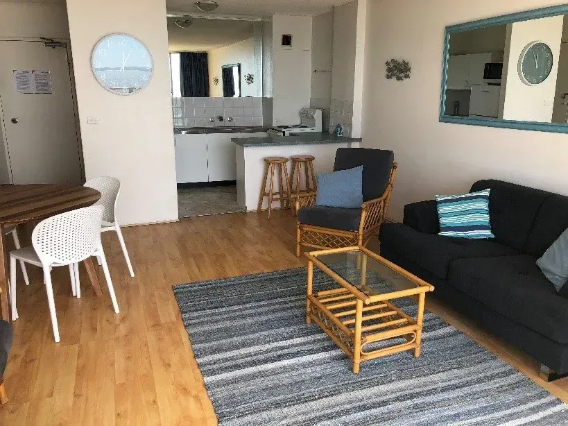 Living area with couch, rug, table, chairs, and kitchen in the background. Wooden floor, light walls, and ocean-themed decor.