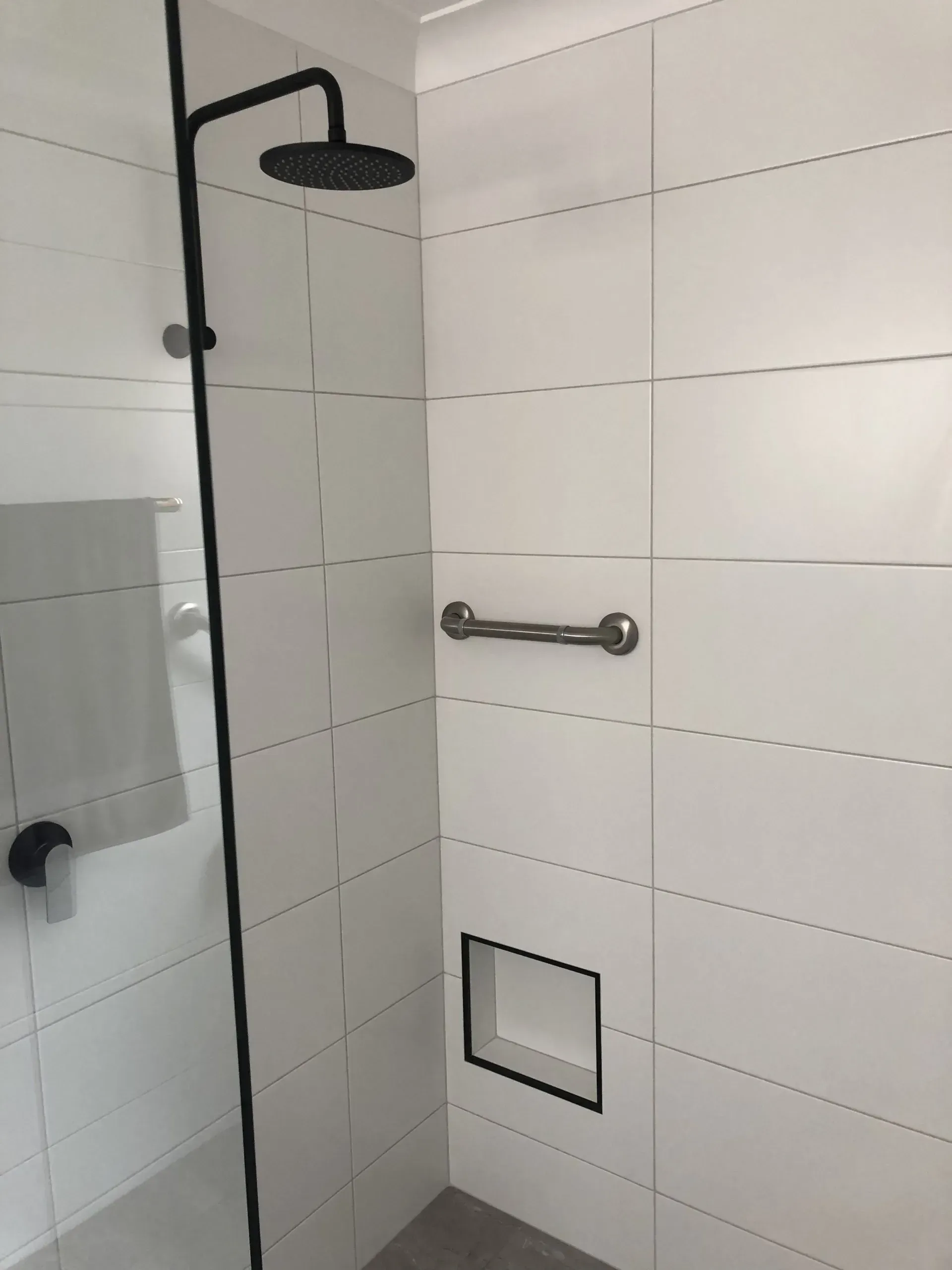 Shower with a black showerhead and glass enclosure, white tiled walls, a grab bar, and a recessed shelf.