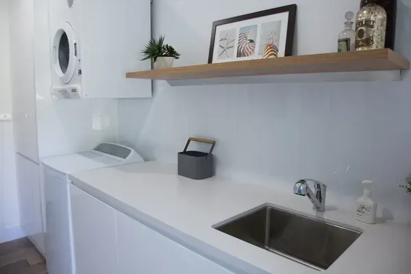 Modern white laundry room with a washing machine, sink, and shelf with decor.