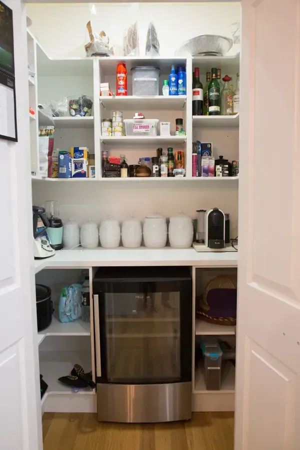 Pantry with white shelves holding food, wine cooler, and jars. White doors frame the entrance.