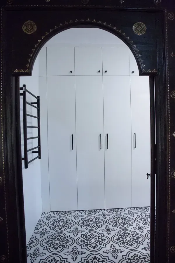 A dark, arched doorway frames white cabinets, a black towel rack, and patterned black and white flooring.