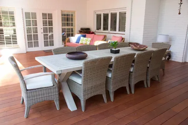Large outdoor dining table with woven chairs on a wooden deck, with a sofa in the background.
