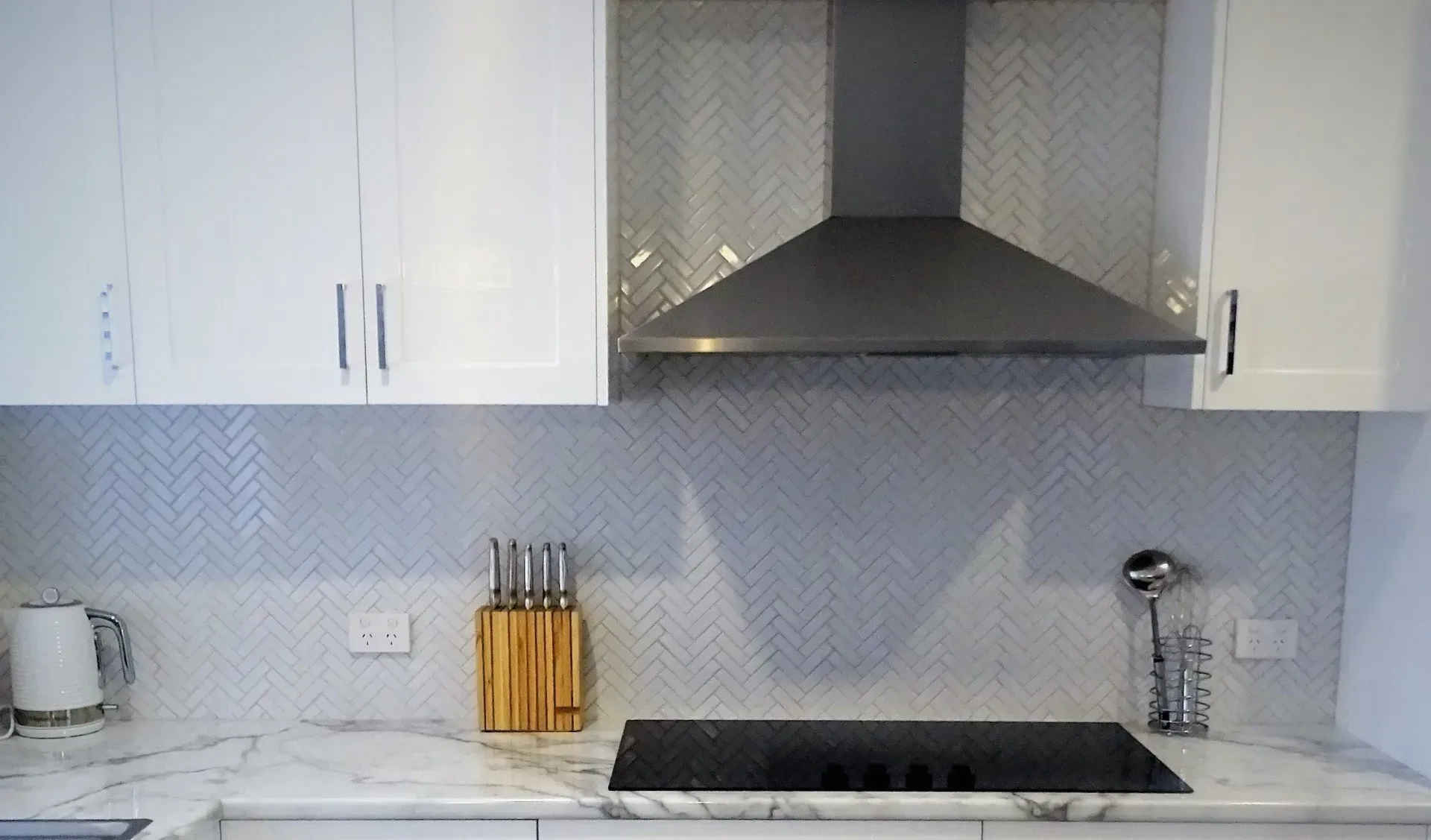 White kitchen with range hood, backsplash, cabinets, countertop, stovetop, knife set, and kettle.
