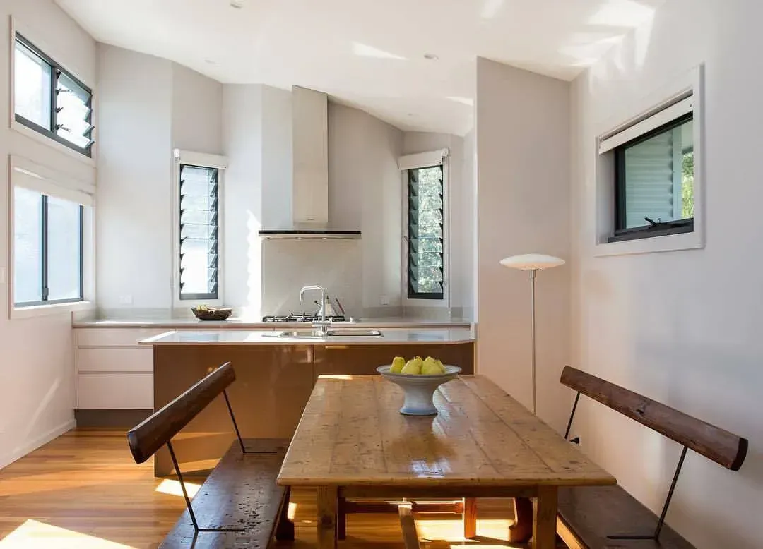 A kitchen with a wooden table and benches, light-colored walls, and dark-framed windows.