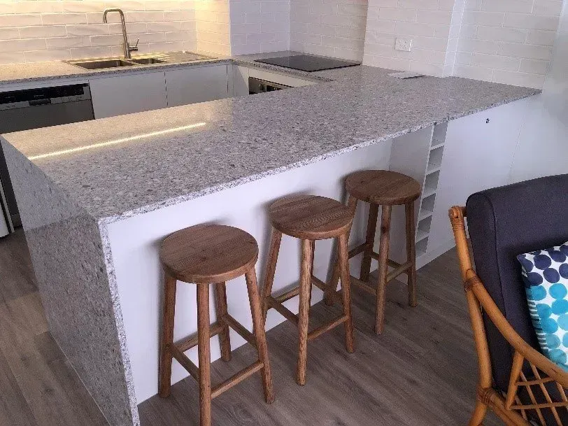 A modern kitchen with a granite countertop, three wooden stools, and built-in wine rack.