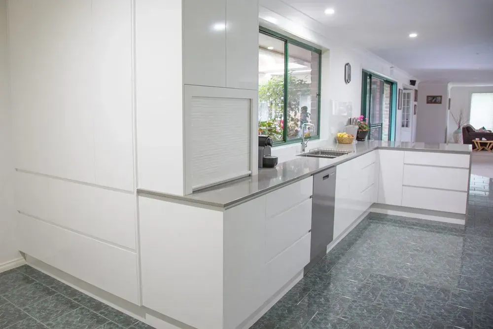 Modern white kitchen with gray countertops, green-tiled floor, and a window overlooking a garden.