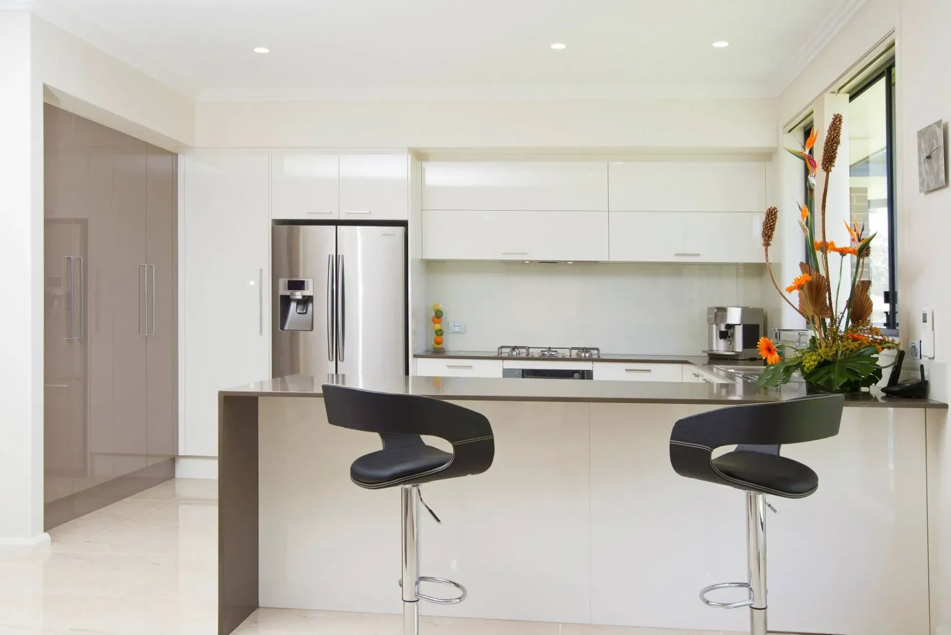 Modern white kitchen with stainless steel appliances, a gray countertop, and two black bar stools.
