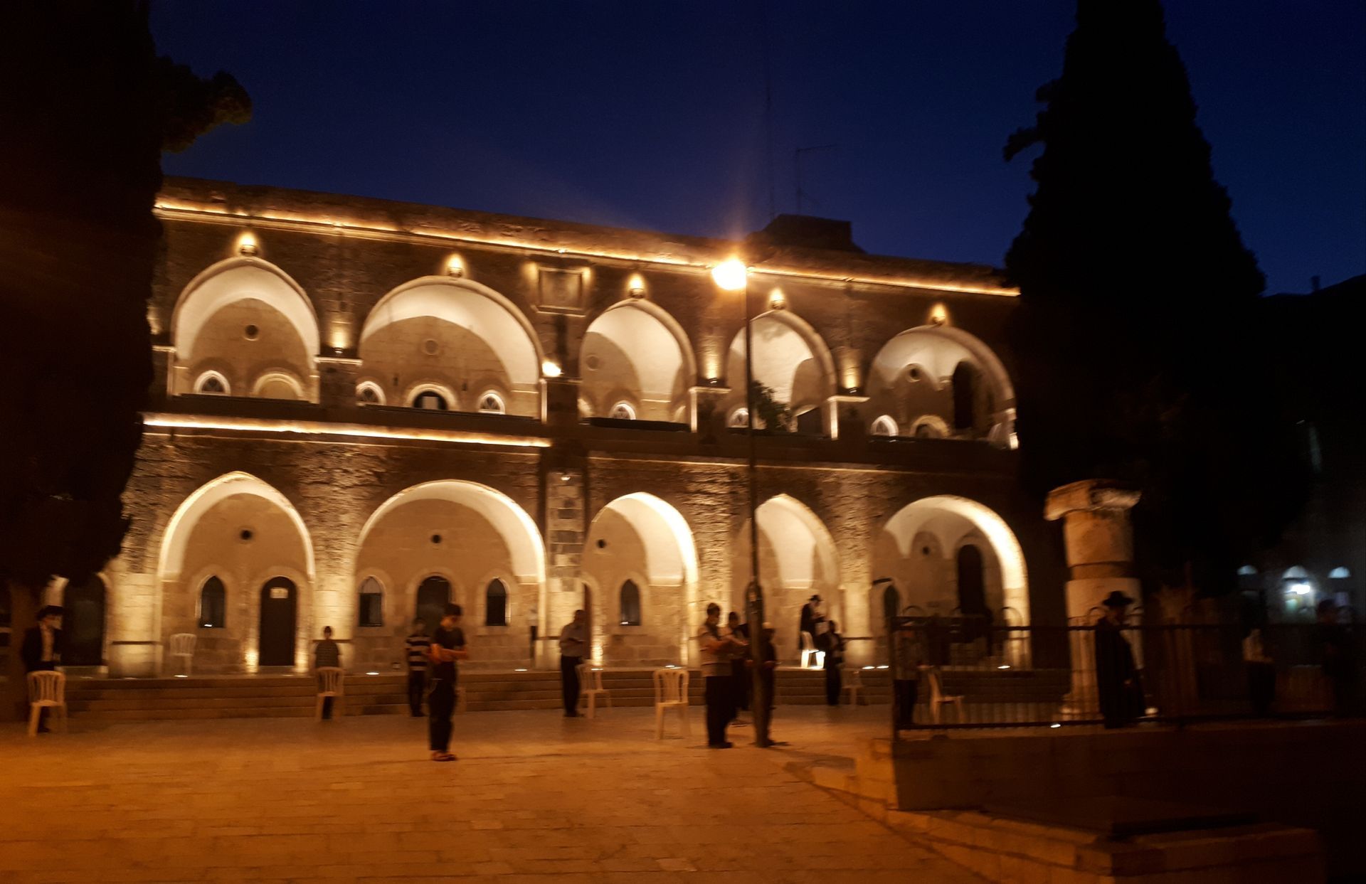 Night view of a stone building with arched windows and lights, people in the courtyard.