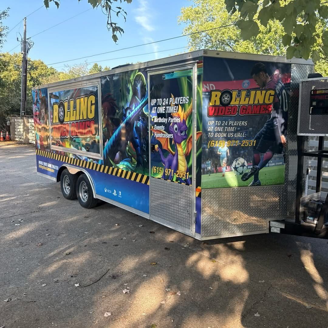 A Rolling Video Games trailer parked on a gravel lot, featuring colorful graphics of game characters and a soccer player.