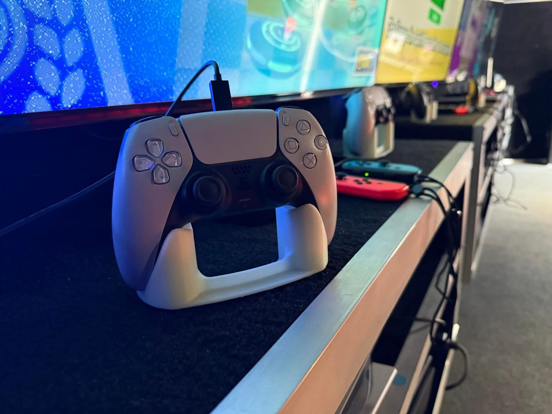 A white PlayStation 5 DualSense controller rests in a charging dock on a black surface in front of large gaming screens.