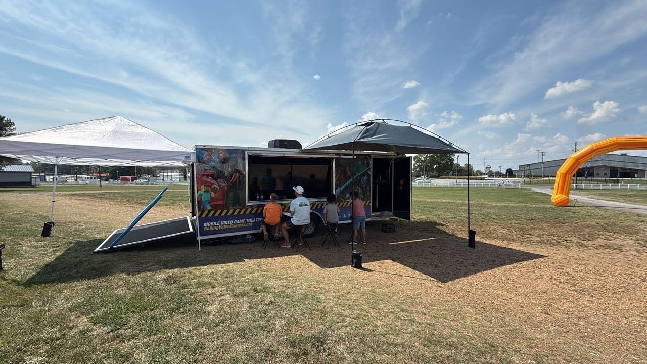 A mobile gaming trailer with an open side awning parked in a grassy field under a sunny sky, with people gathered inside.