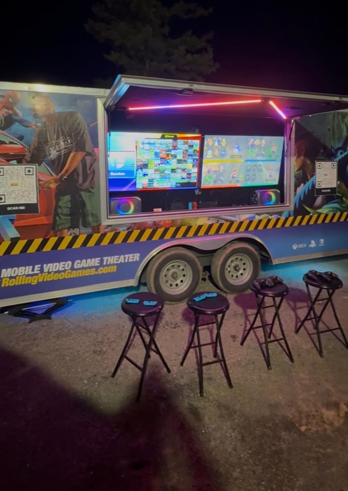 A mobile video game theater trailer parked at night with four bar stools, two exterior screens, and blue under-lighting.