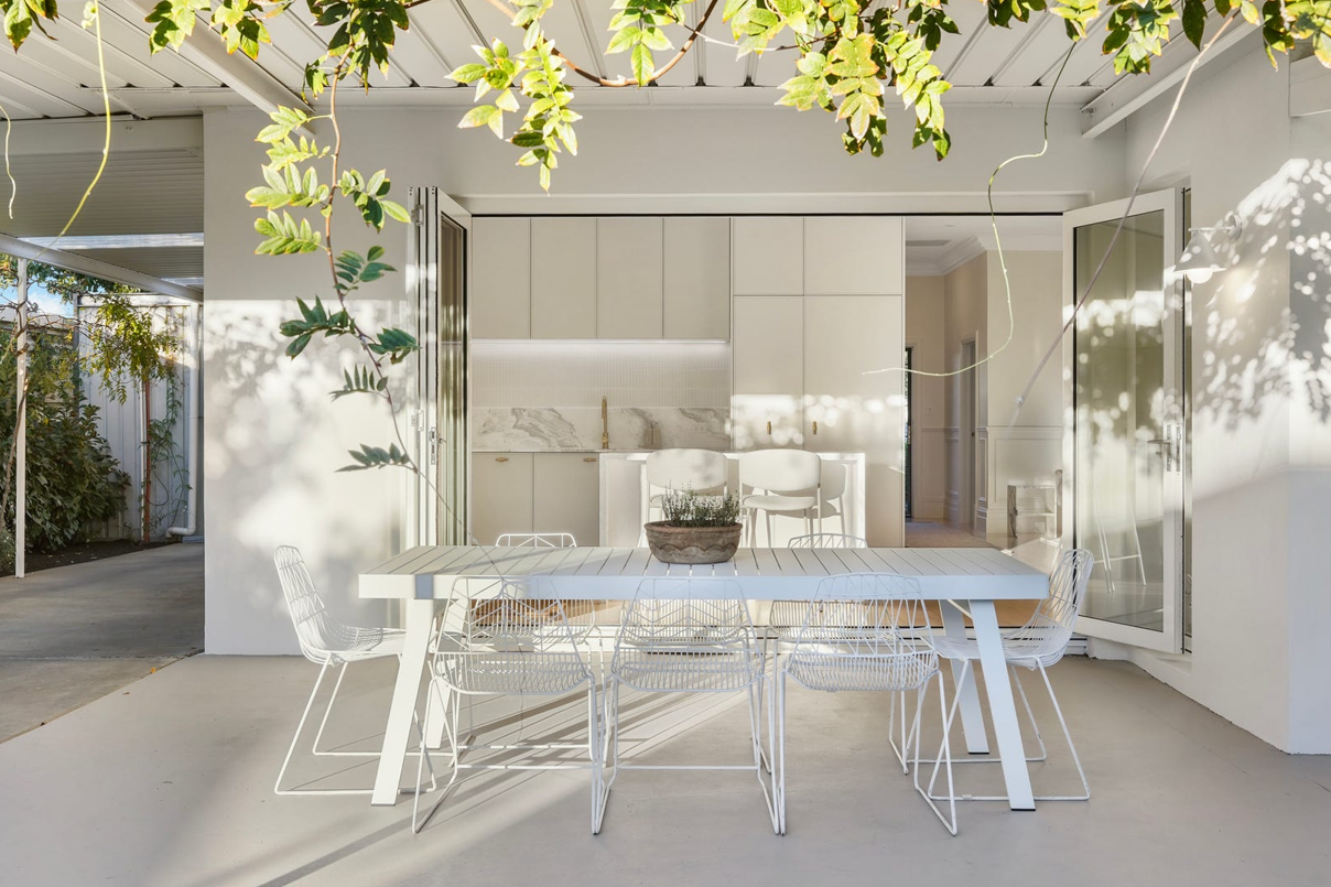 A white dining table and chairs on a patio with a kitchen in the background.