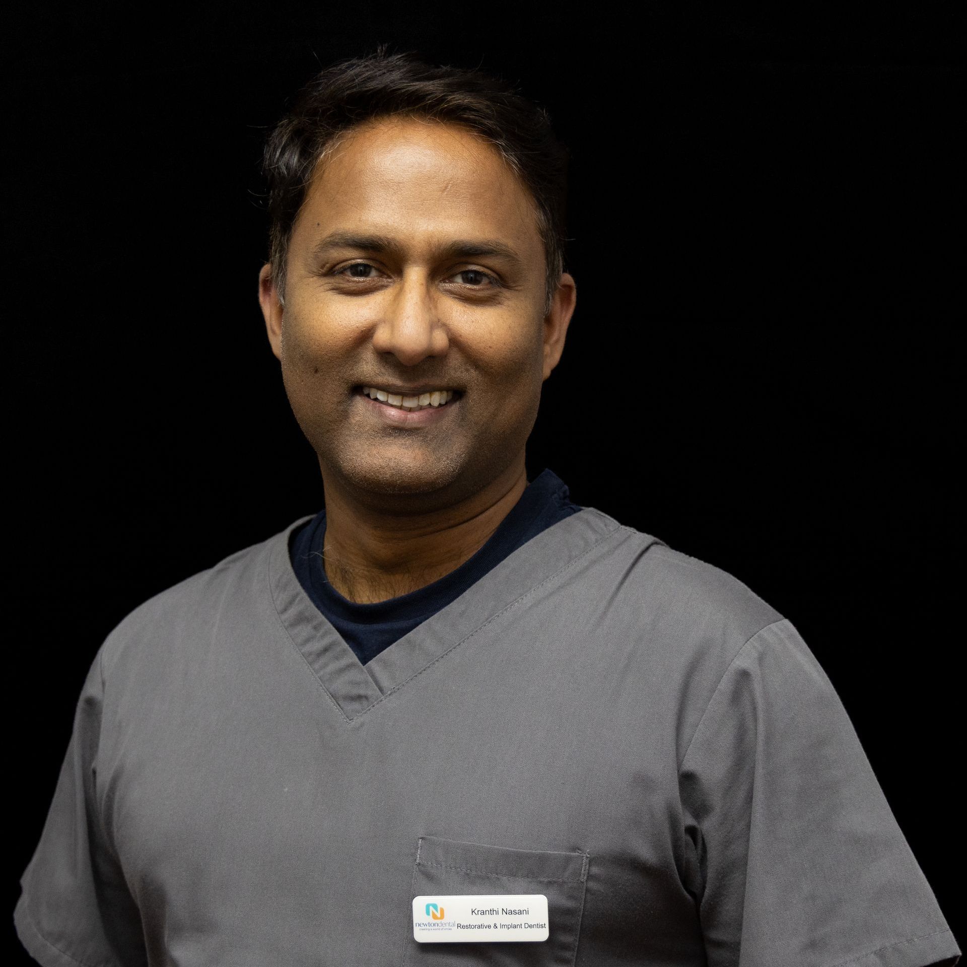 A man in a scrub top with a name tag on his pocket smiles for the camera.