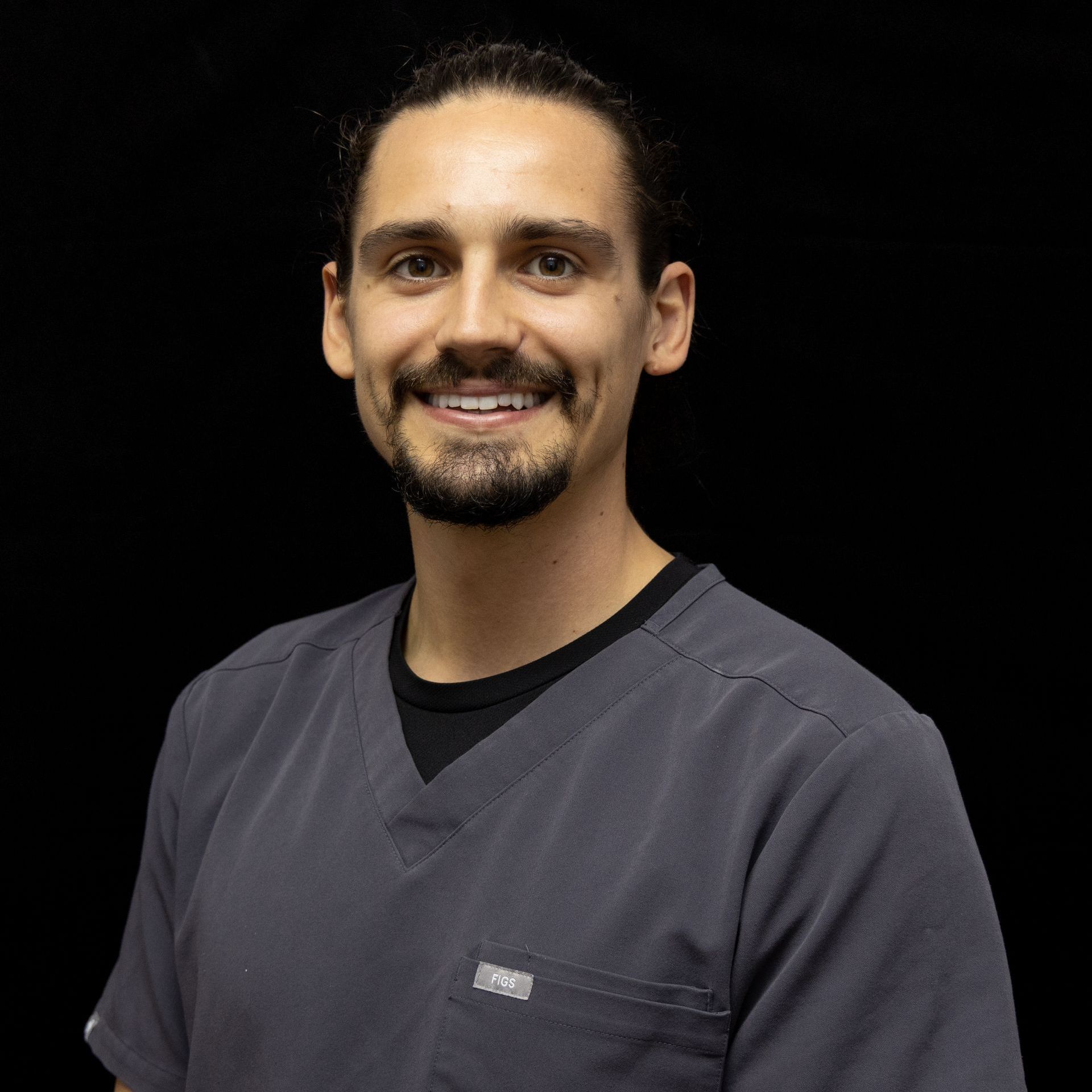 A man with a beard is wearing a scrub top and smiling for the camera.