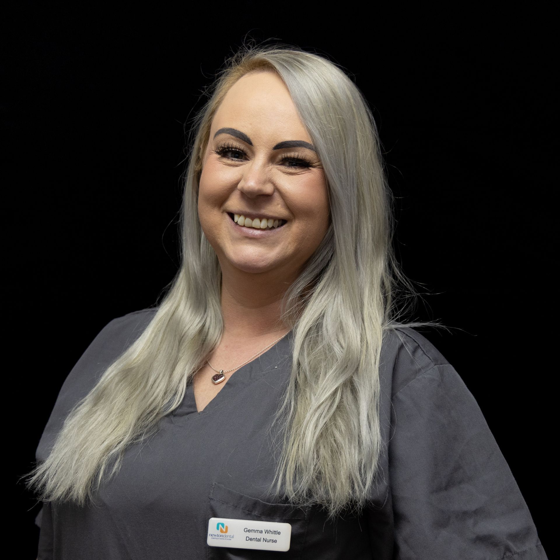 A woman with long blonde hair is wearing a scrub and smiling.