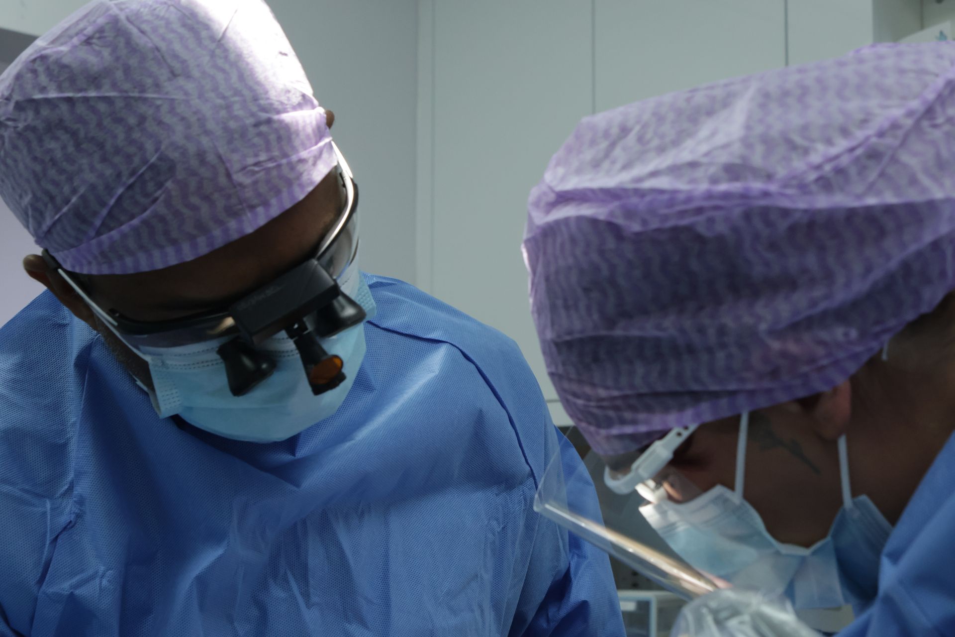 Two surgeons in blue scrubs and caps, operating for dental implants. One wears magnification glasses.