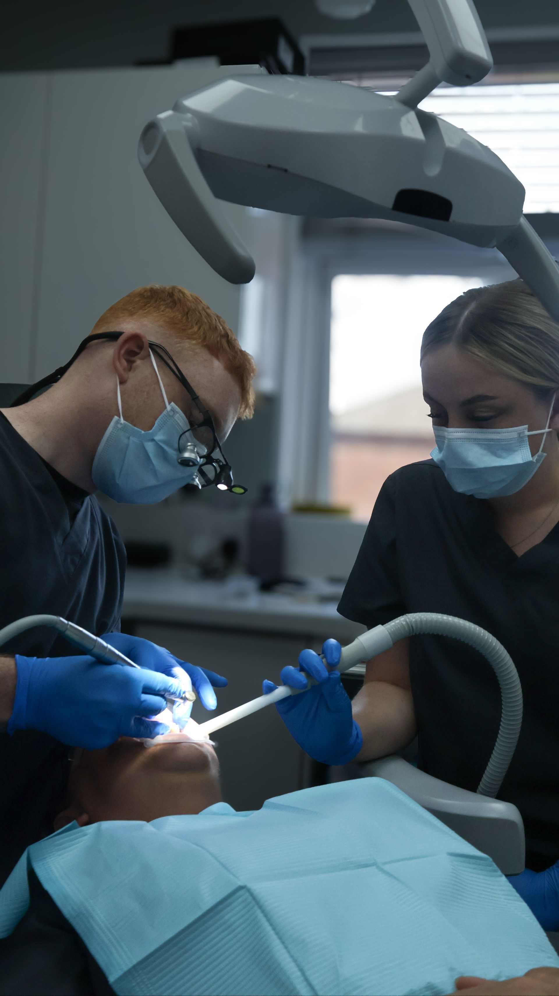 a dentist and a dental nurse, in a clinical environment, performing dental treatment. General dentistry. The dentist is wearing loop glasses. 
