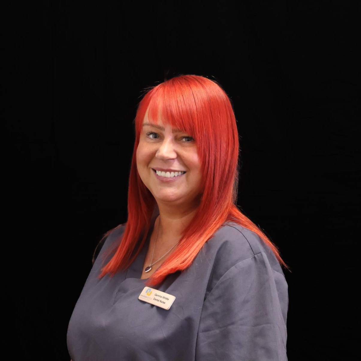 A woman with long red hair is wearing a scrub and smiling.