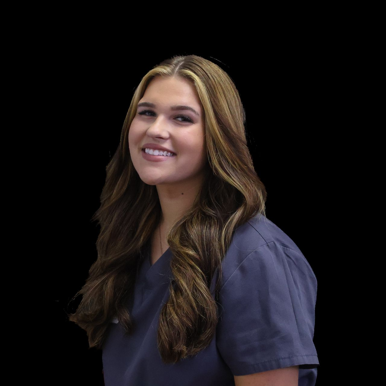 A woman with long brunette hair is wearing a gray scrub top and smiling.