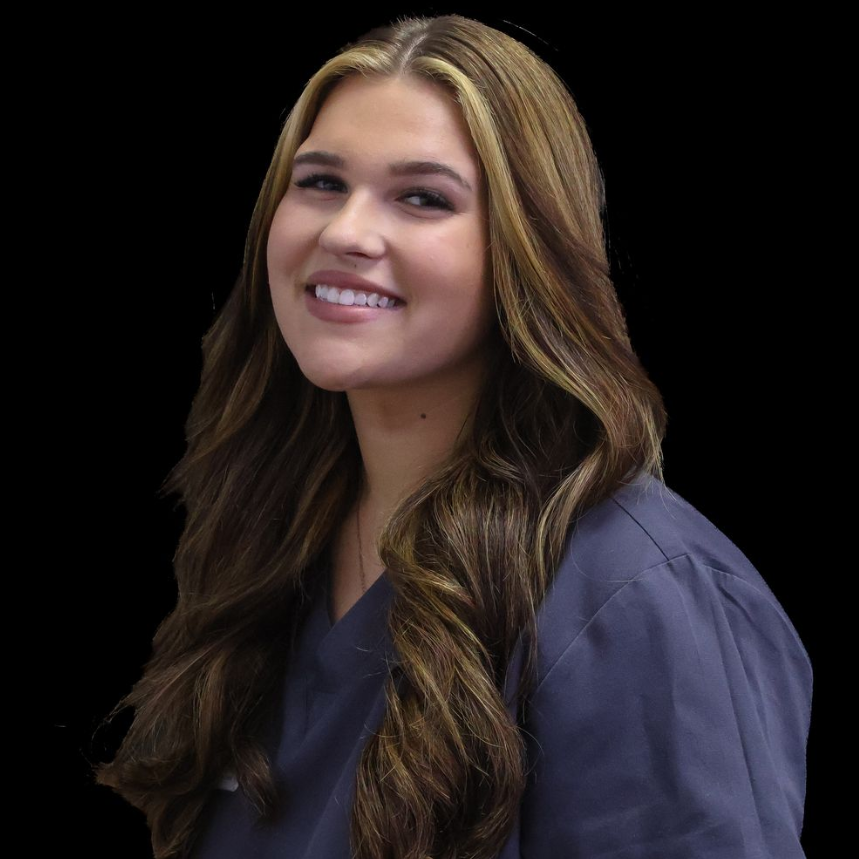 A woman with long brunette hair is wearing a gray scrub top and smiling.