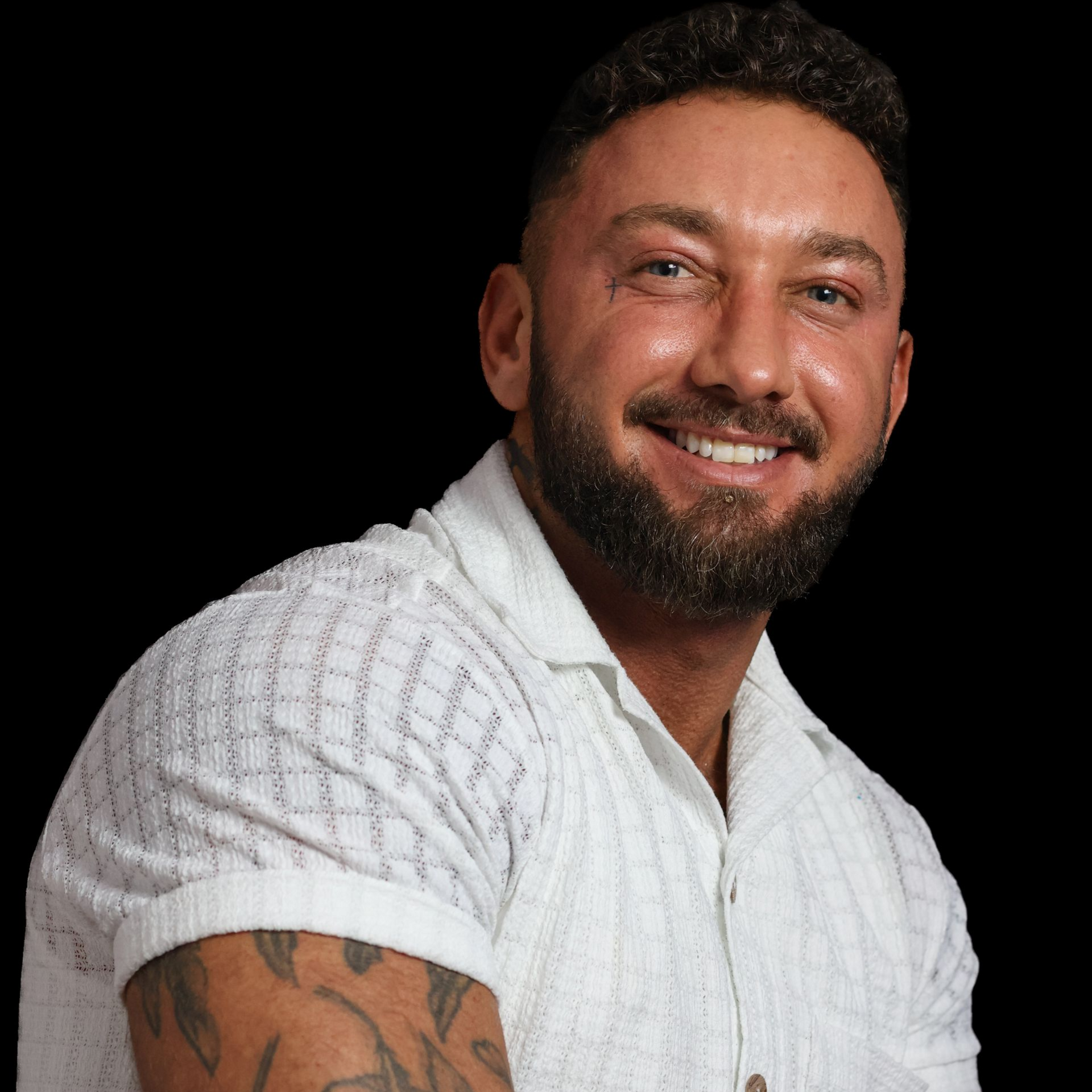 A man with a beard and tattoos is wearing a white shirt.