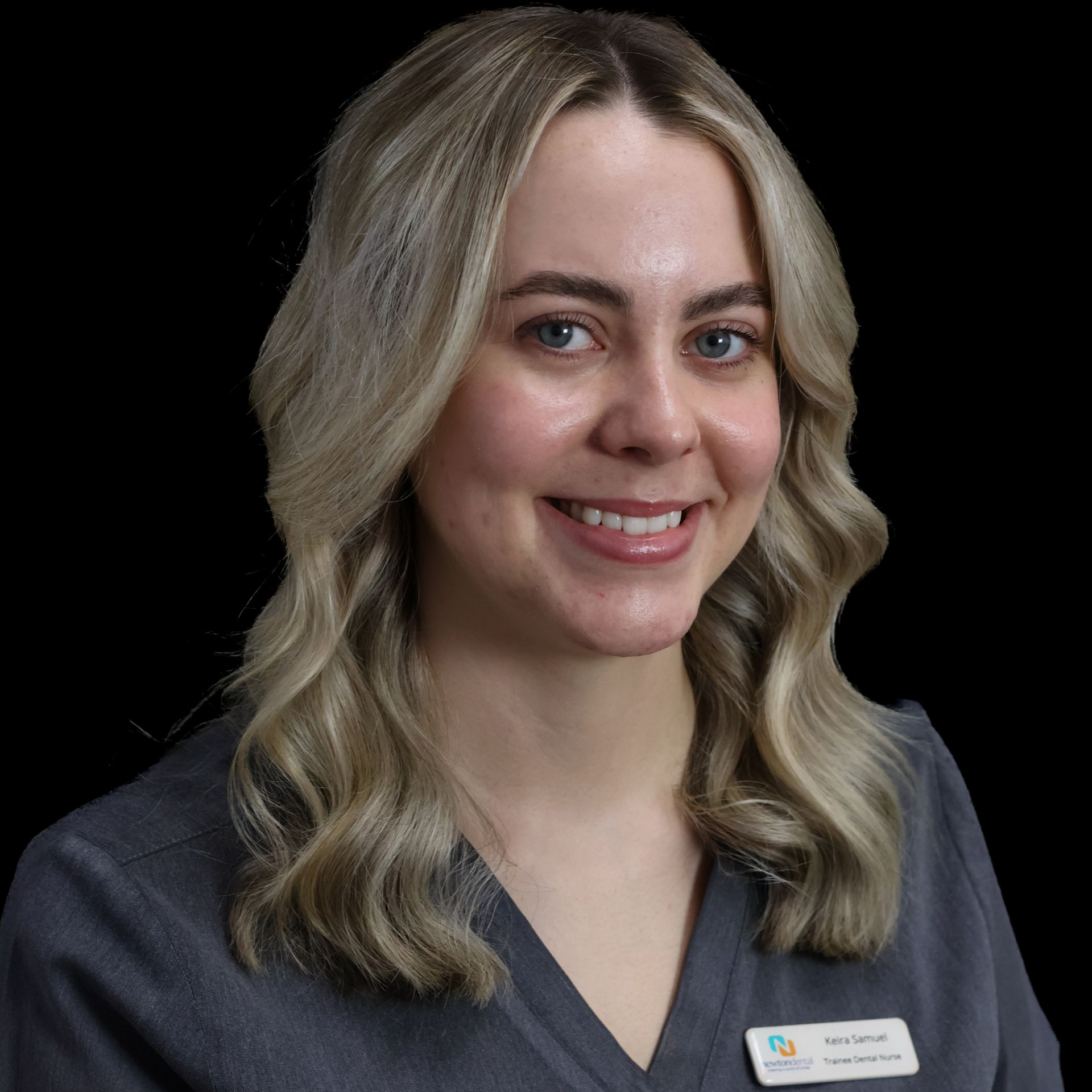 A woman with long blonde hair is wearing a grey scrub and smiling for the camera in a clinical setting.