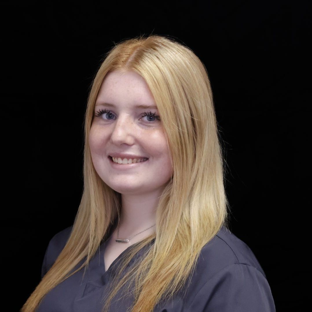 A woman with long blonde hair is wearing a gray scrub top and smiling.
