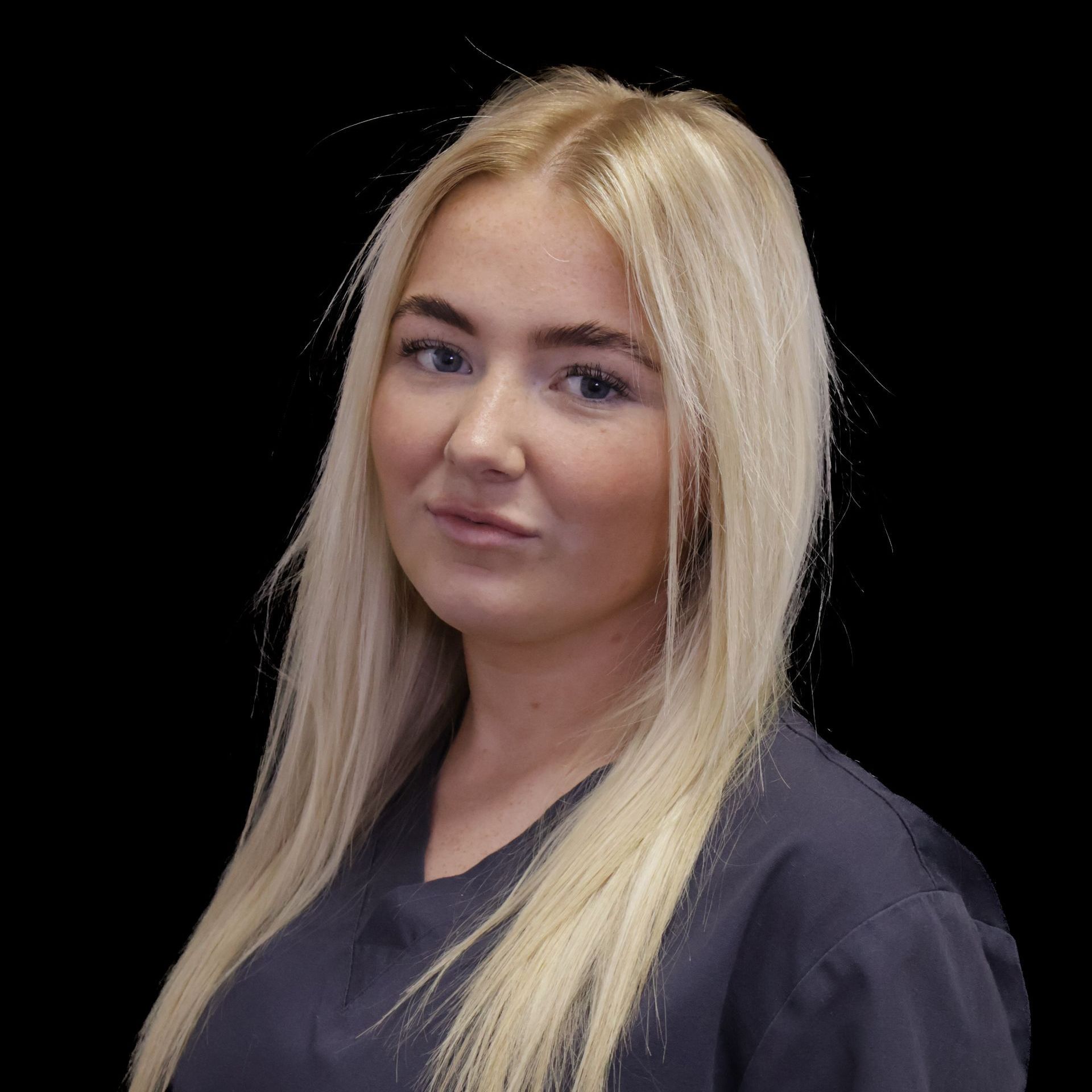 A woman with long blonde hair is wearing a grey scrub and smiling for the camera in a clinical setting.