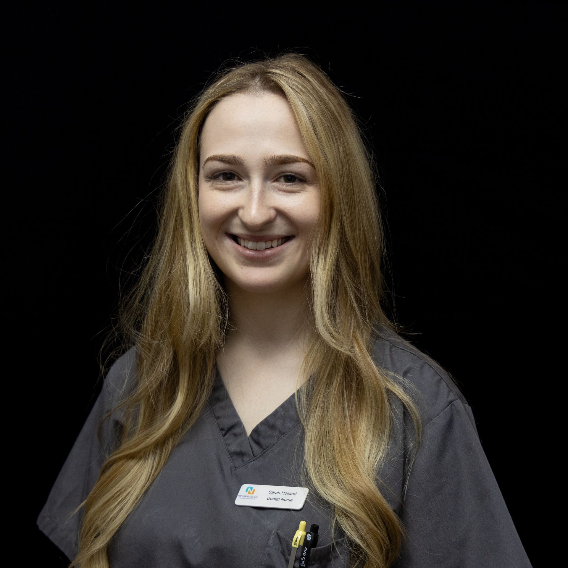 A woman with long blonde hair is wearing a grey scrub top and smiling.