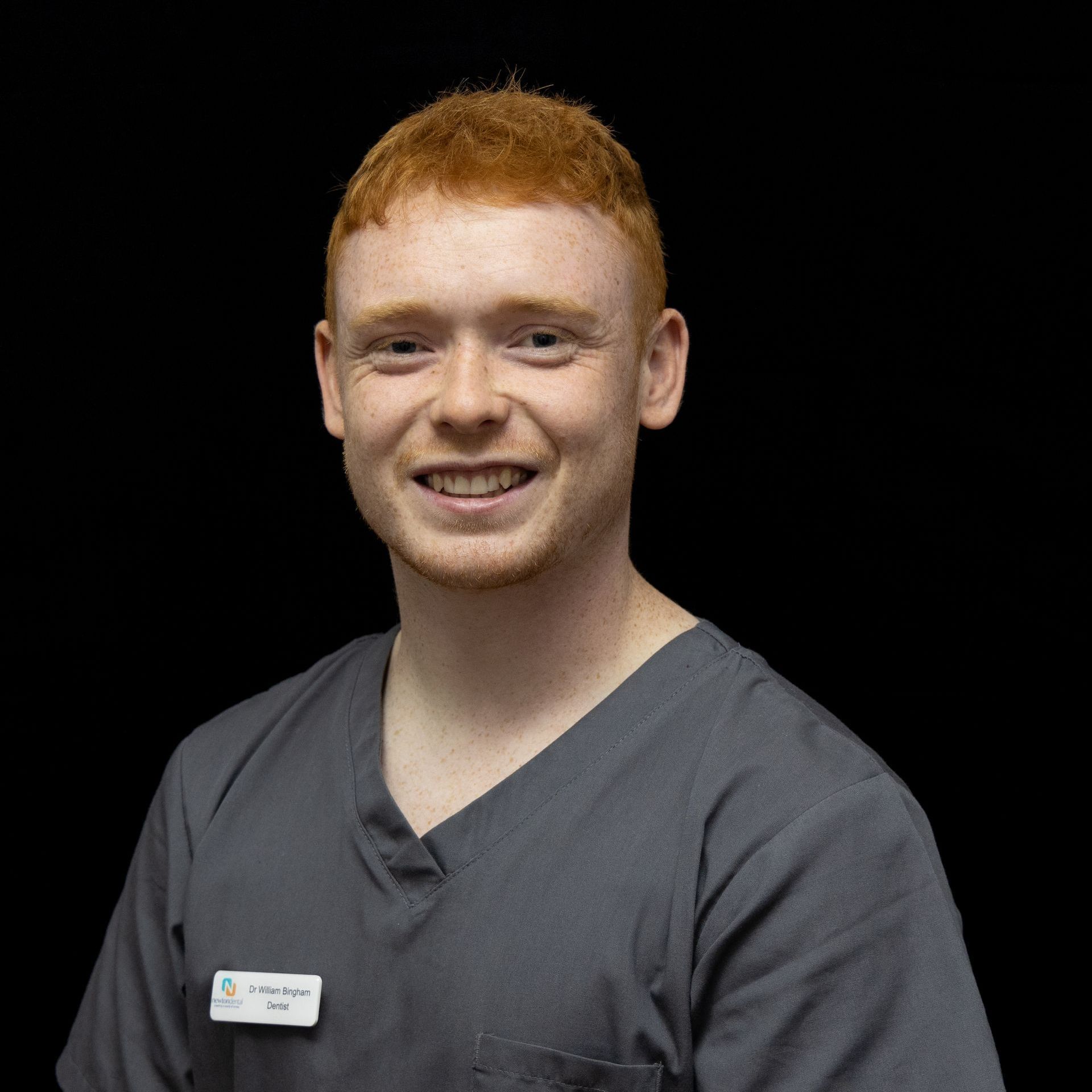 A man with red hair is wearing a gray scrub top and smiling.