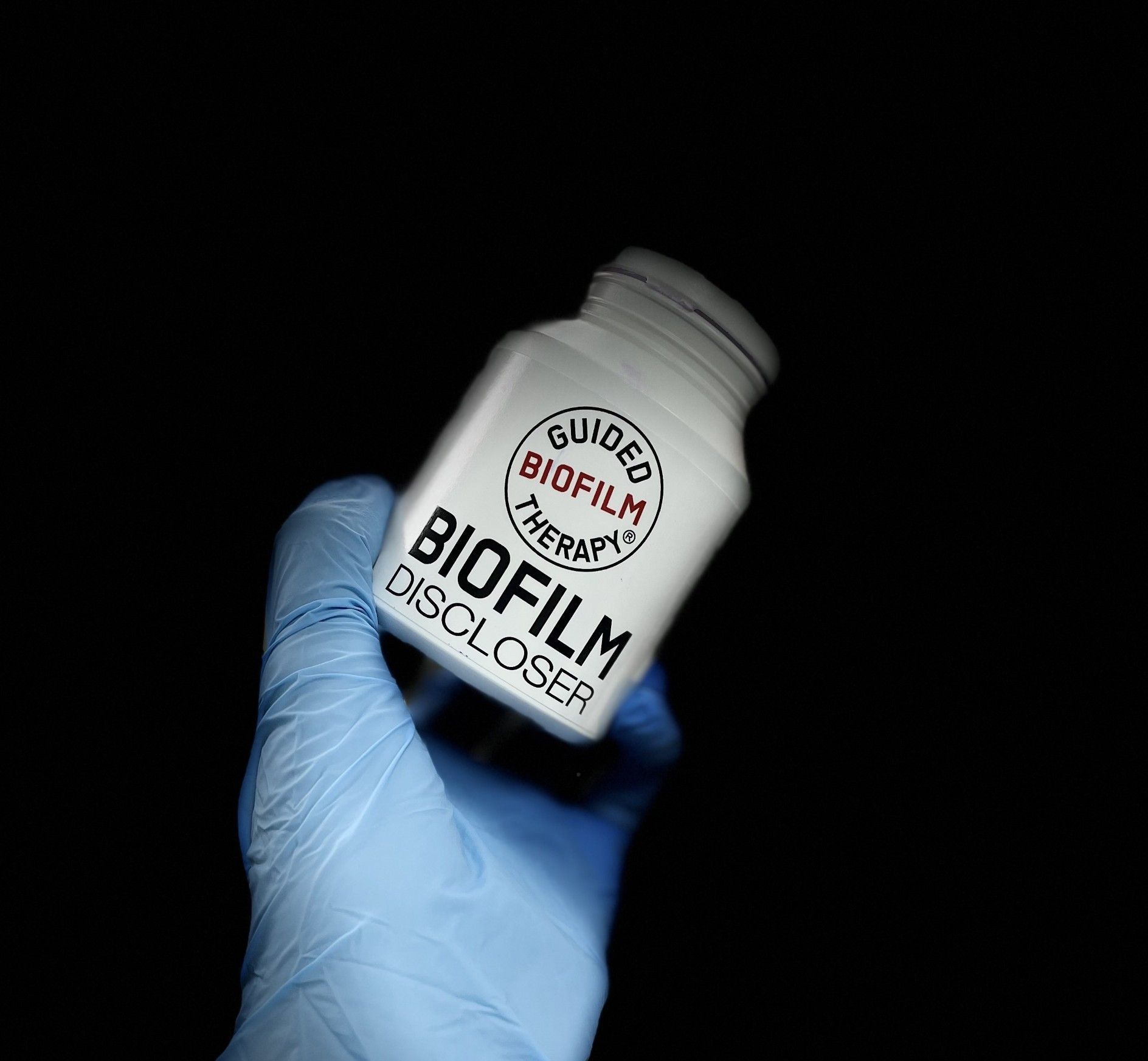 A hand wearing blue gloves is holding a bottle of biofilm discloser for airflow treatment