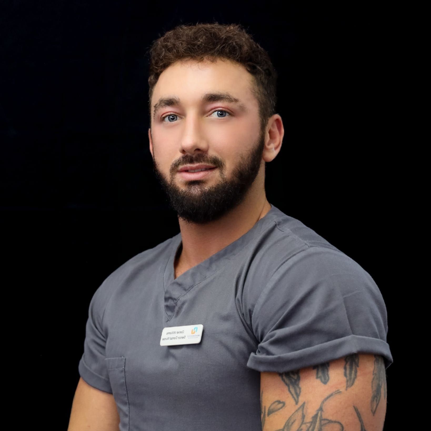 A man with a beard and tattoos is wearing a grey scrub top.