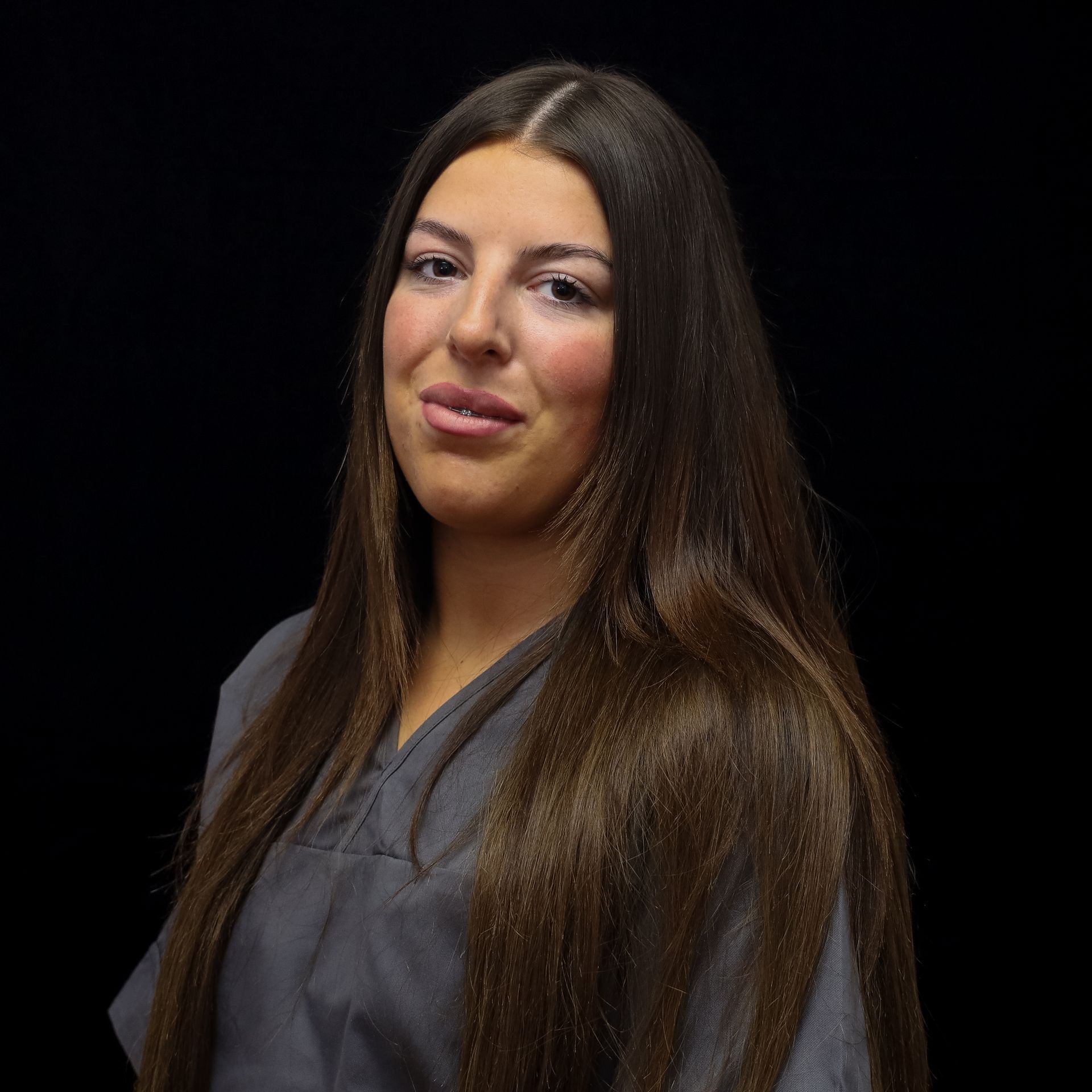 A woman with long hair is wearing a grey scrub and smiling for the camera.