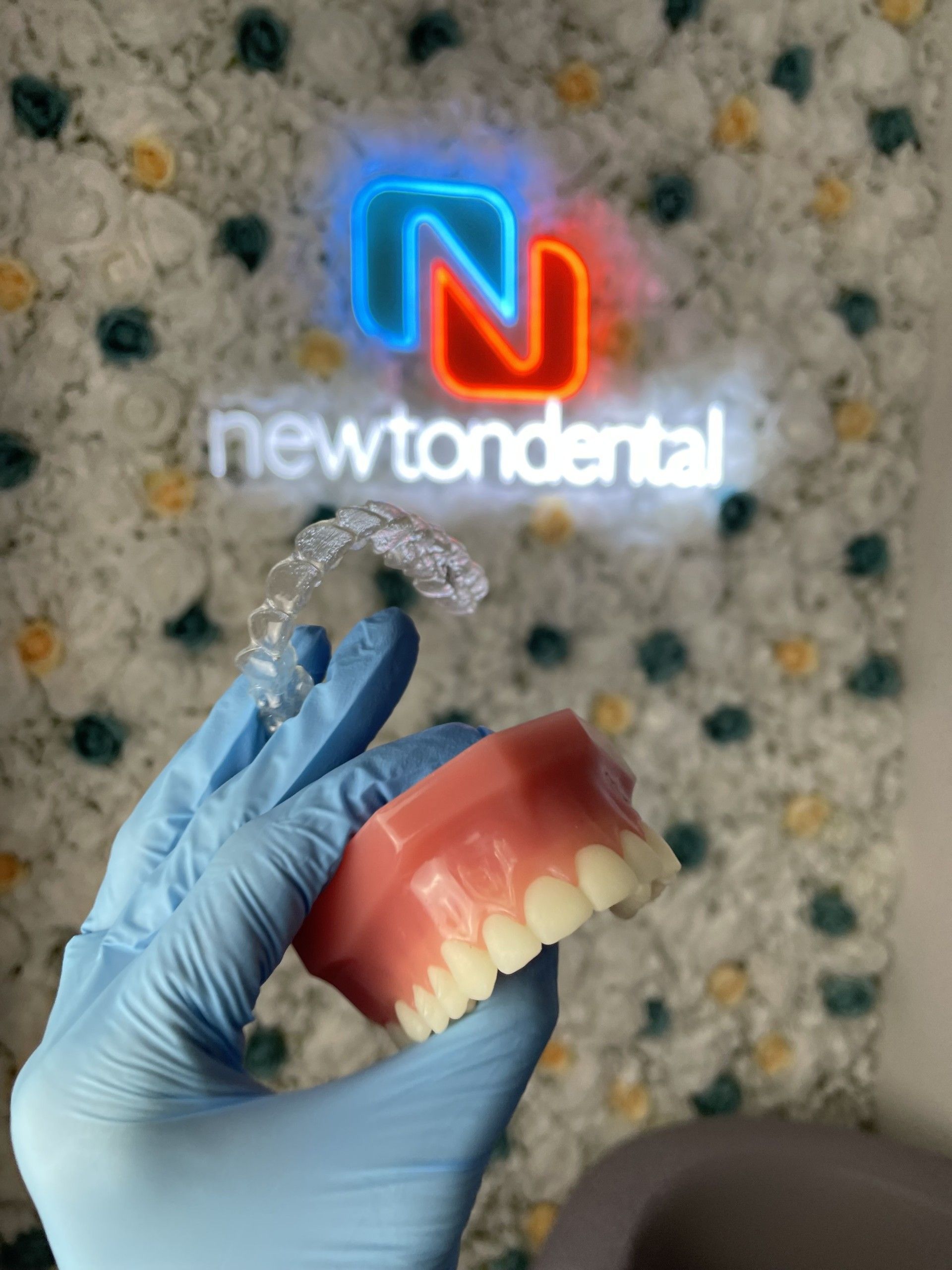 A person is holding a model of teeth in front of a neon sign that says newtondental. Clear and invisible aligners.