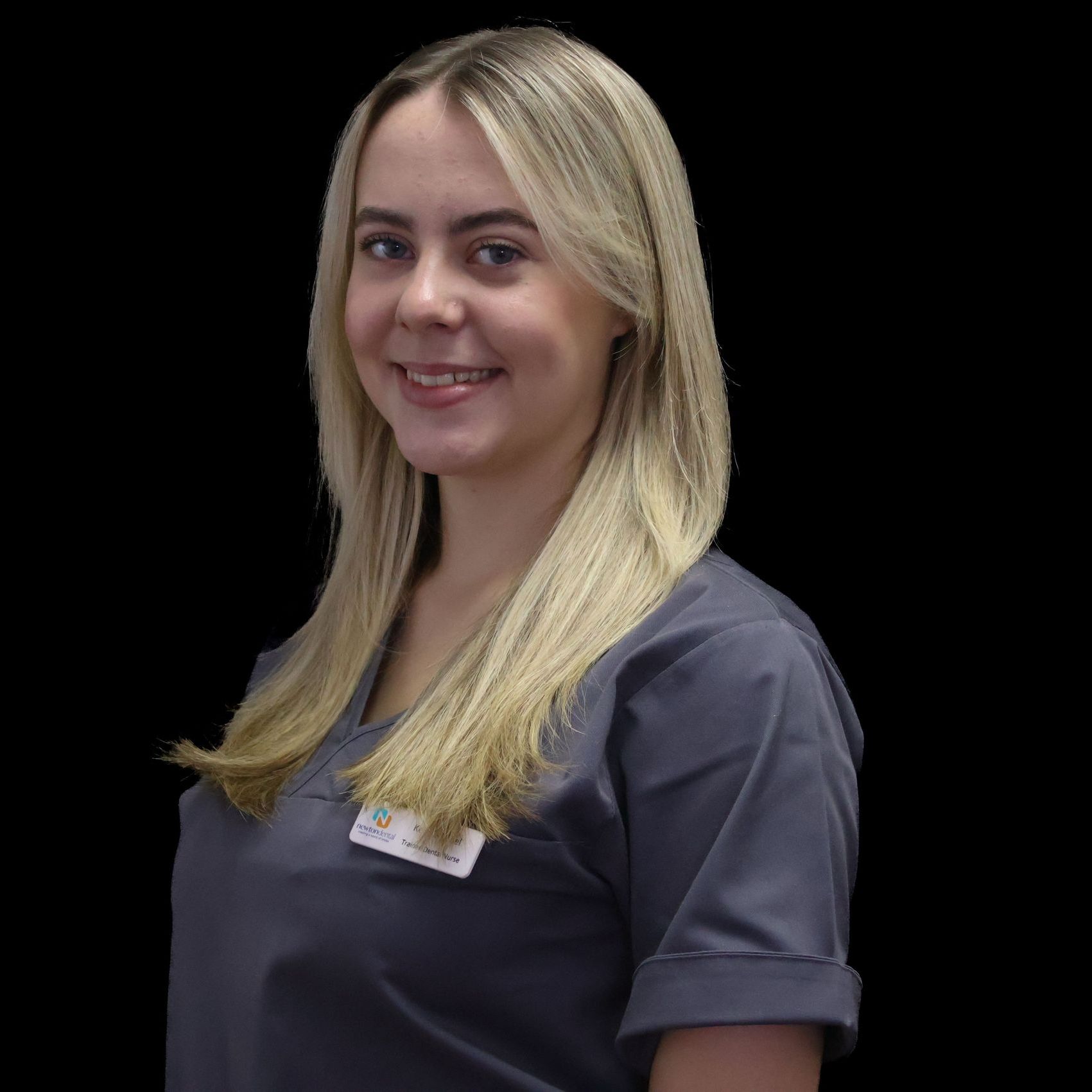 A woman with long blonde hair is wearing a gray scrub top and smiling.