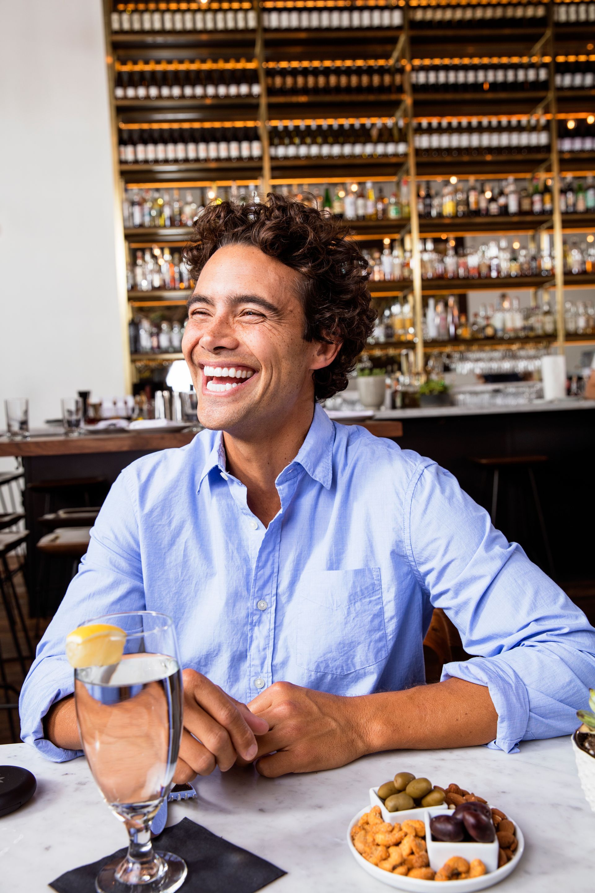 Man laughing at a bar, sitting at a table with a drink and snacks with beautiful straight teeth. 