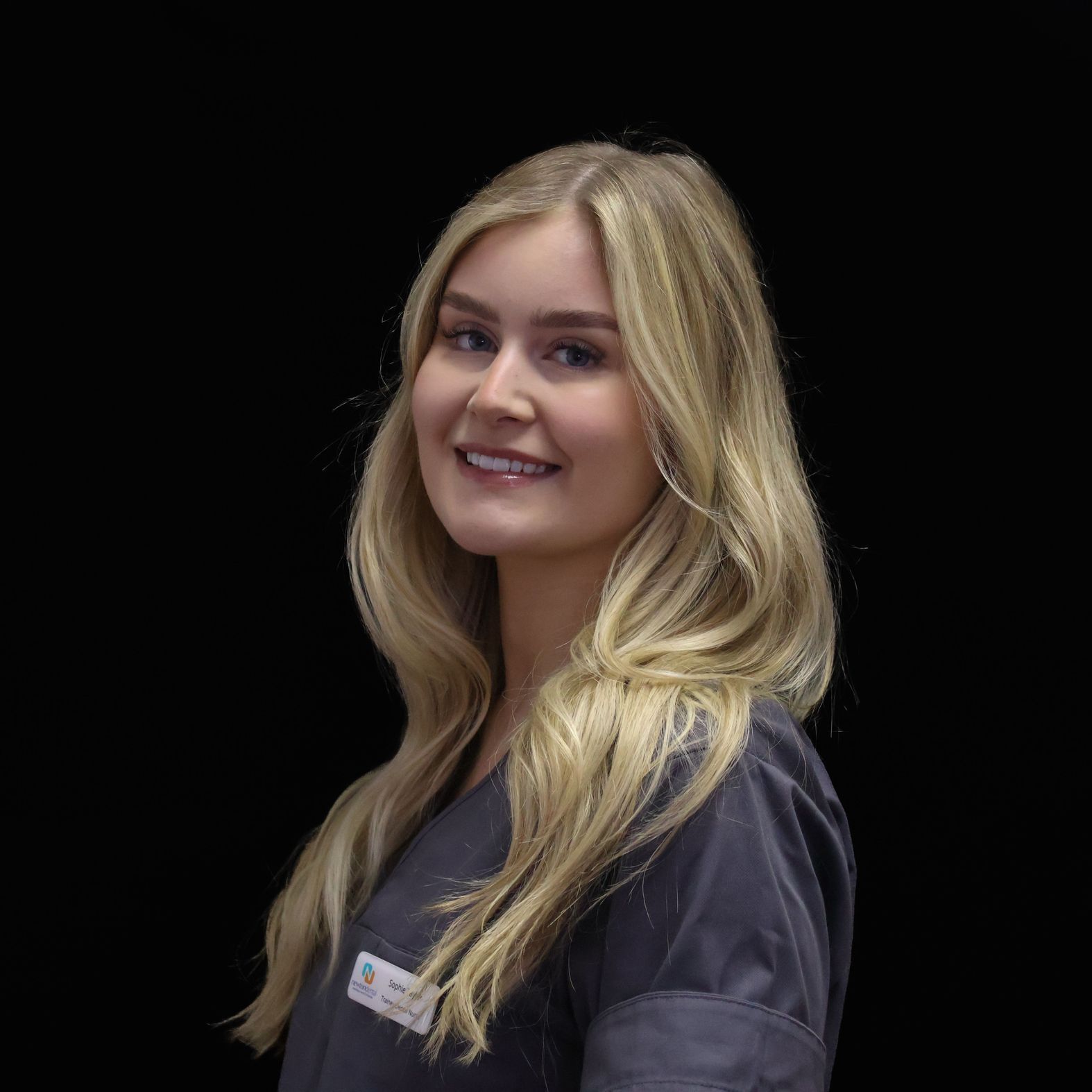 A woman with long blonde hair is wearing a grey scrub top and smiling.