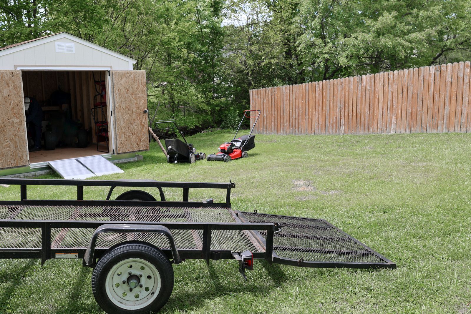 A trailer is parked in the grass in front of a shed.