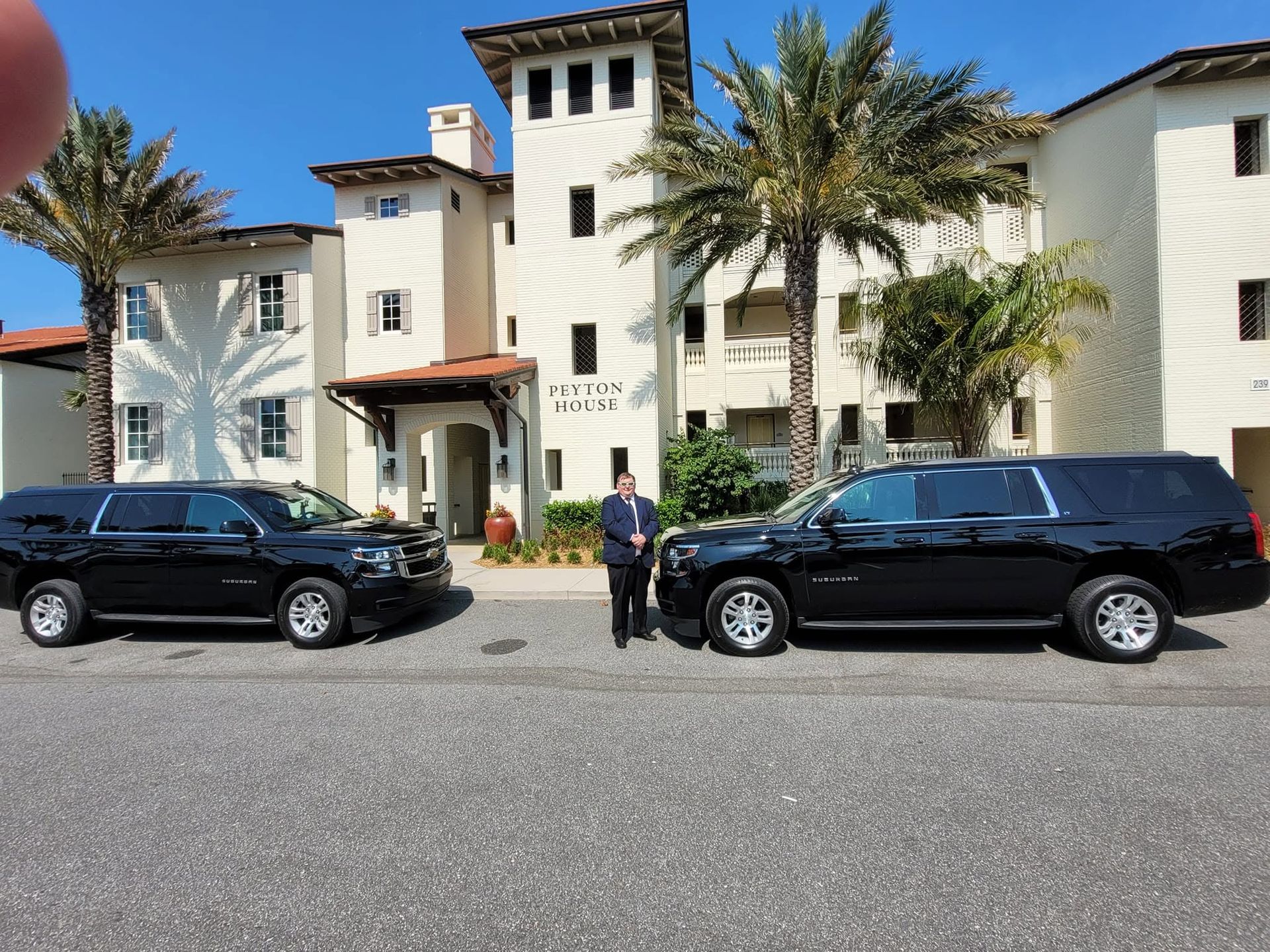 Two black SUVs parked in front of a white Mediterranean-style building with palm trees