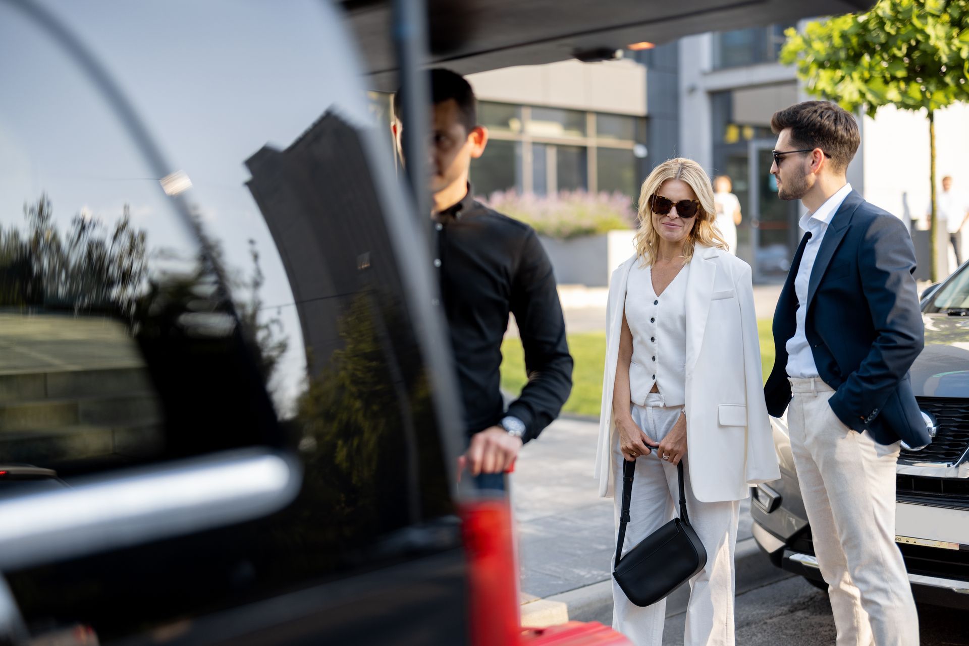 Three people standing beside a black vehicle, with a woman in white and sunglasses in the center.