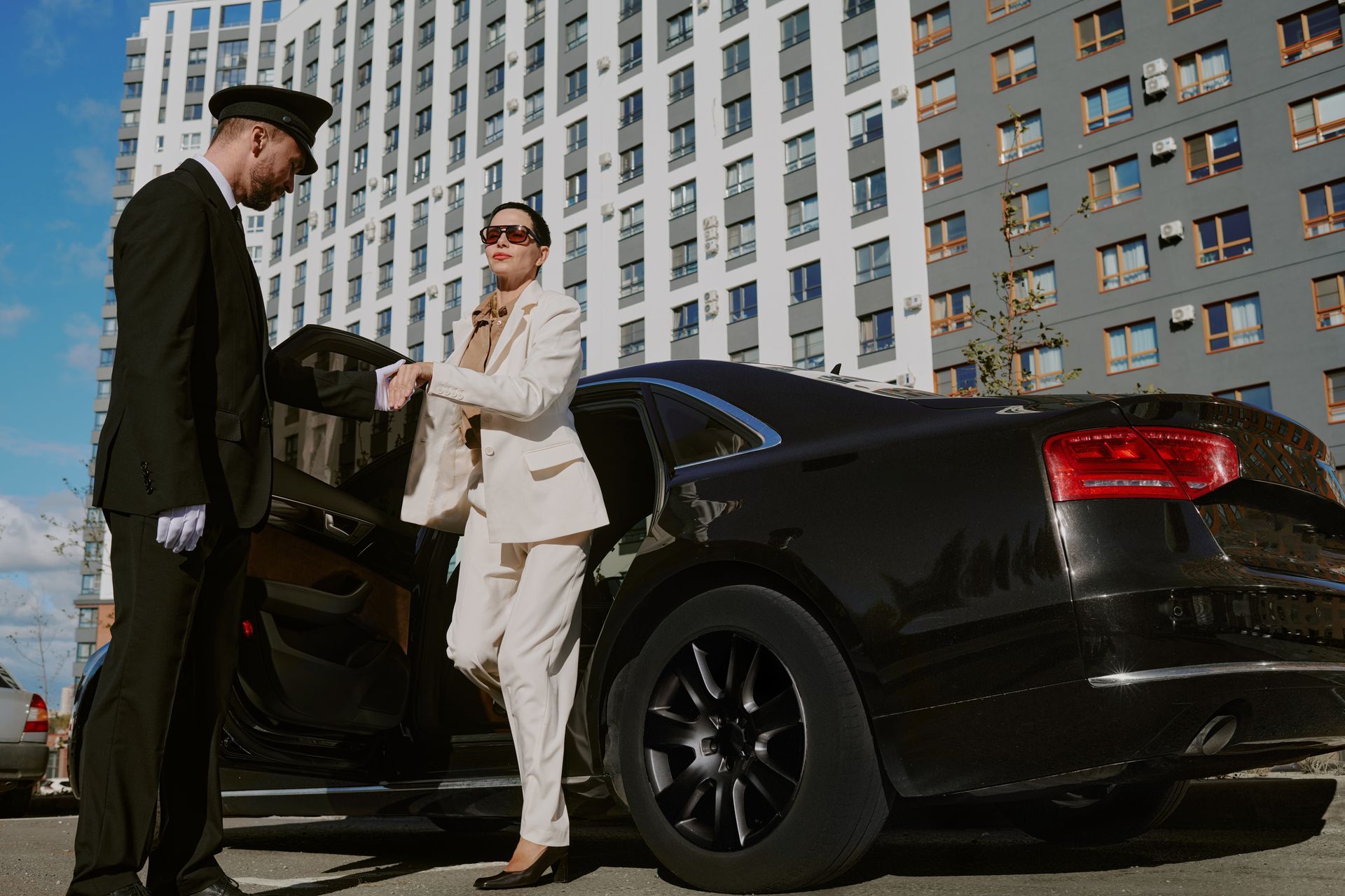 Two men in suits standing by a black sedan with the rear door open on a city street.