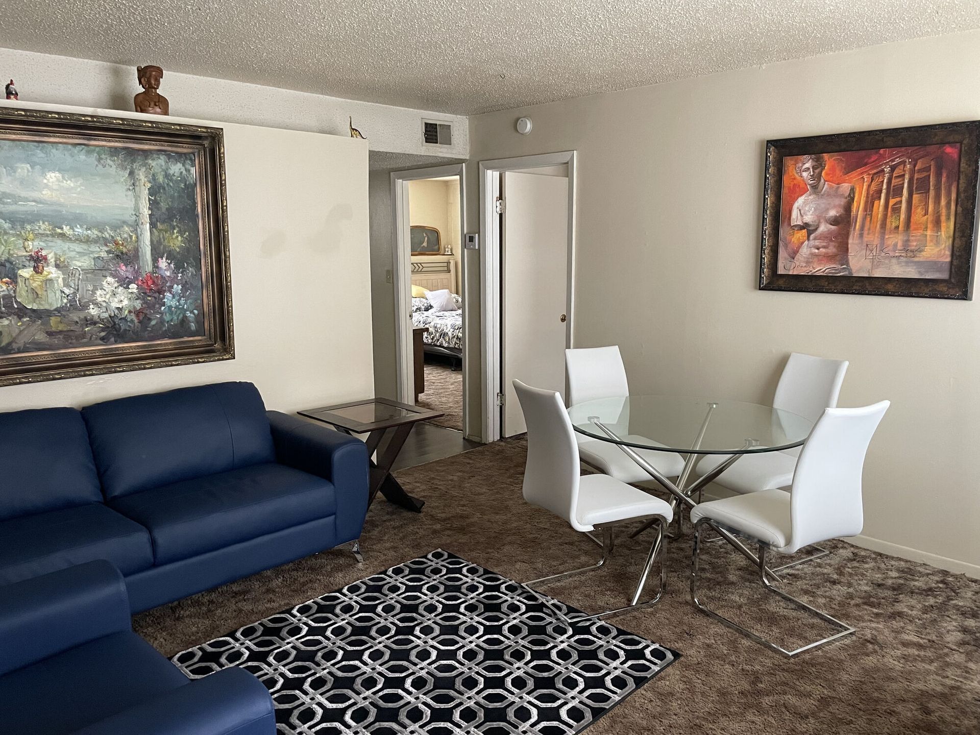 Living room with a blue couch, glass dining table, art, and a view into a bedroom.