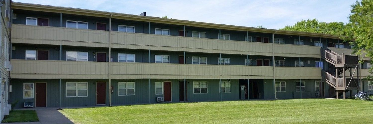 Three-story apartment building with green siding and balconies, set on a grassy lawn with trees in the background.