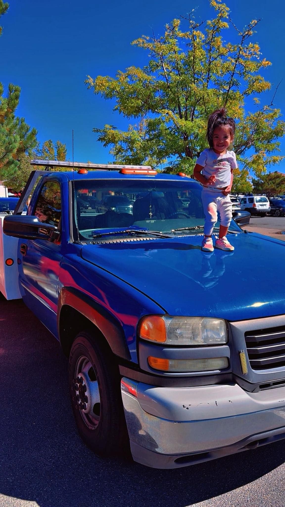 Child standing on the hood of a blue tow truck, with blue sky and trees in the background.
